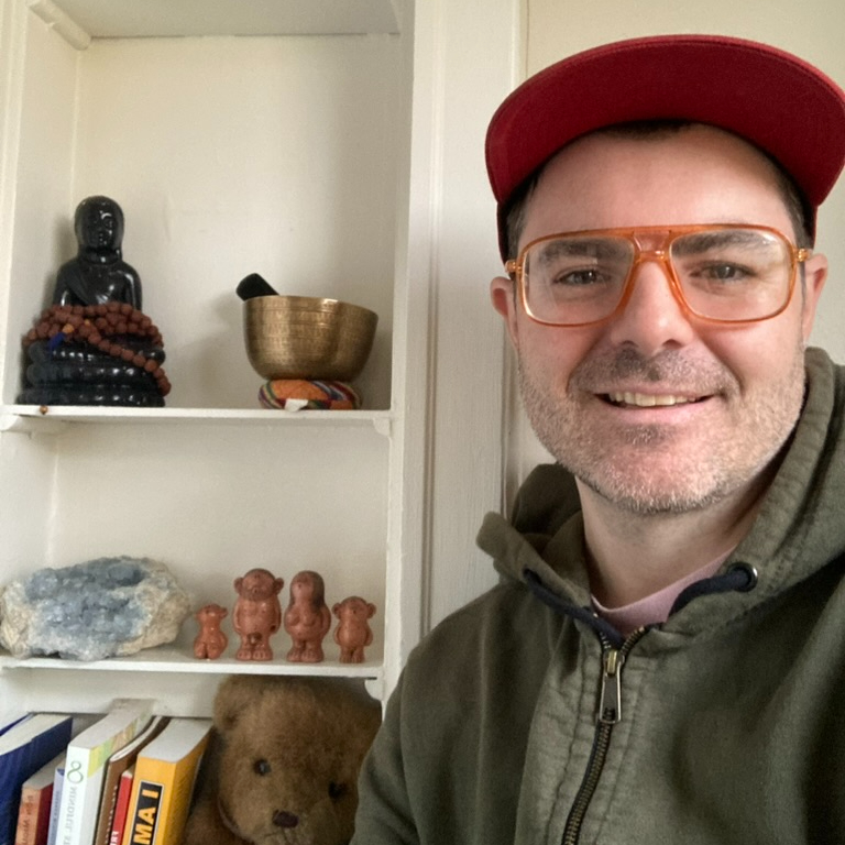 A photo of David in front of a bookshelf with a Buddha statue, singing bowls, and a teddy bear.